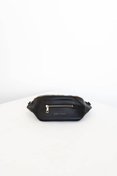 A black belt bag with gold zippers is positioned in the center of a white surface. The bag has two zippered pockets on the front, and the words